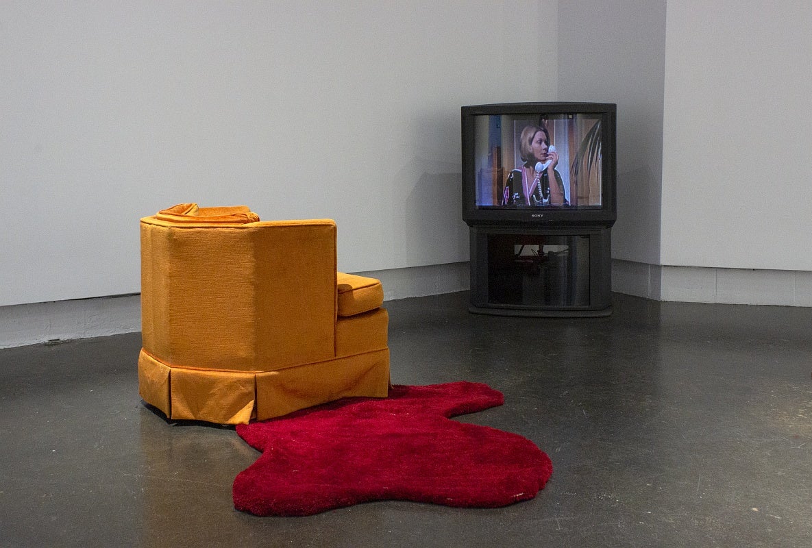 An orange armchair with an irregularly shaped red floor rug that resembles a pool of blood faces a 1980's television set displaying a woman holding a vintage phone and looking over her left shoulder.