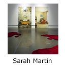 Gallery room with two large photos of room interior and red, irregularly-shaped rugs on the floor, text reads Sarah Martin 