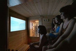 Several smiling women wearing bathing suits and towels are seated in a wood panelled sauna watching a video monitor