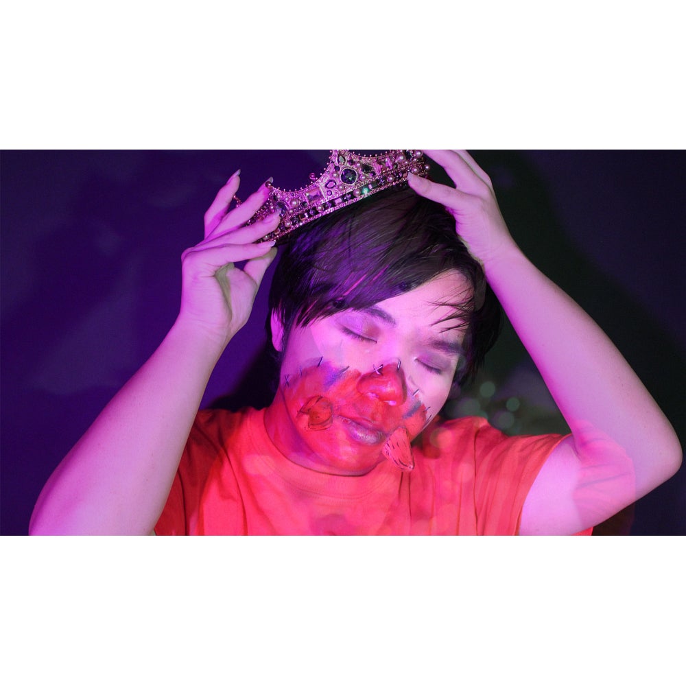 Person, lit by a pink light and with red paint on the lower half of their face, positions a crown on their head.