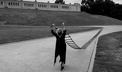 The artist, dressed in matching black dress and face mask, walks along a paved pathway in front of a large water treatment plant, pulling a long nautical rope ladder behind her