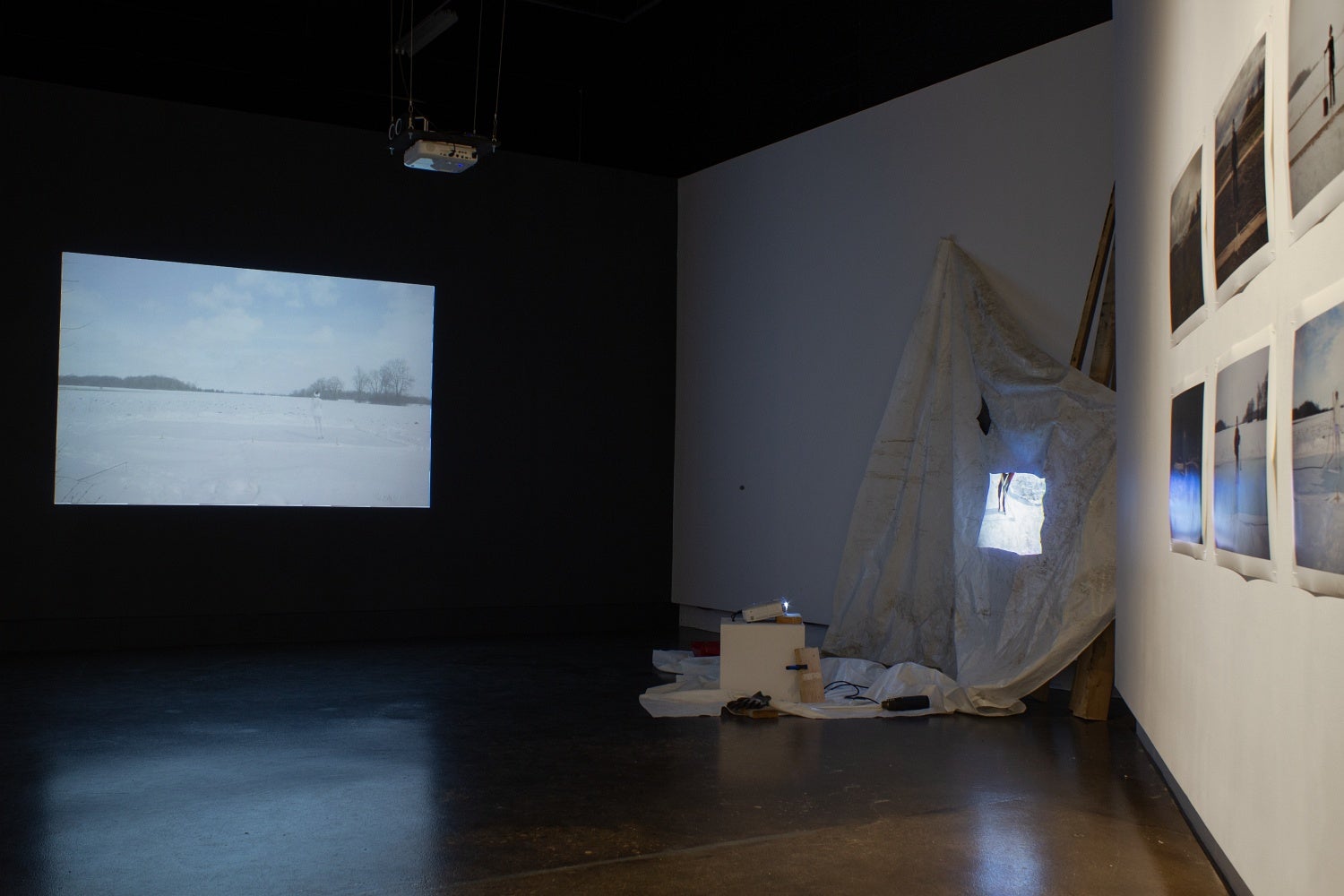 Art gallery with 6 photographs hung in a grid and video projections on a wall and on a crumpled white tarp.