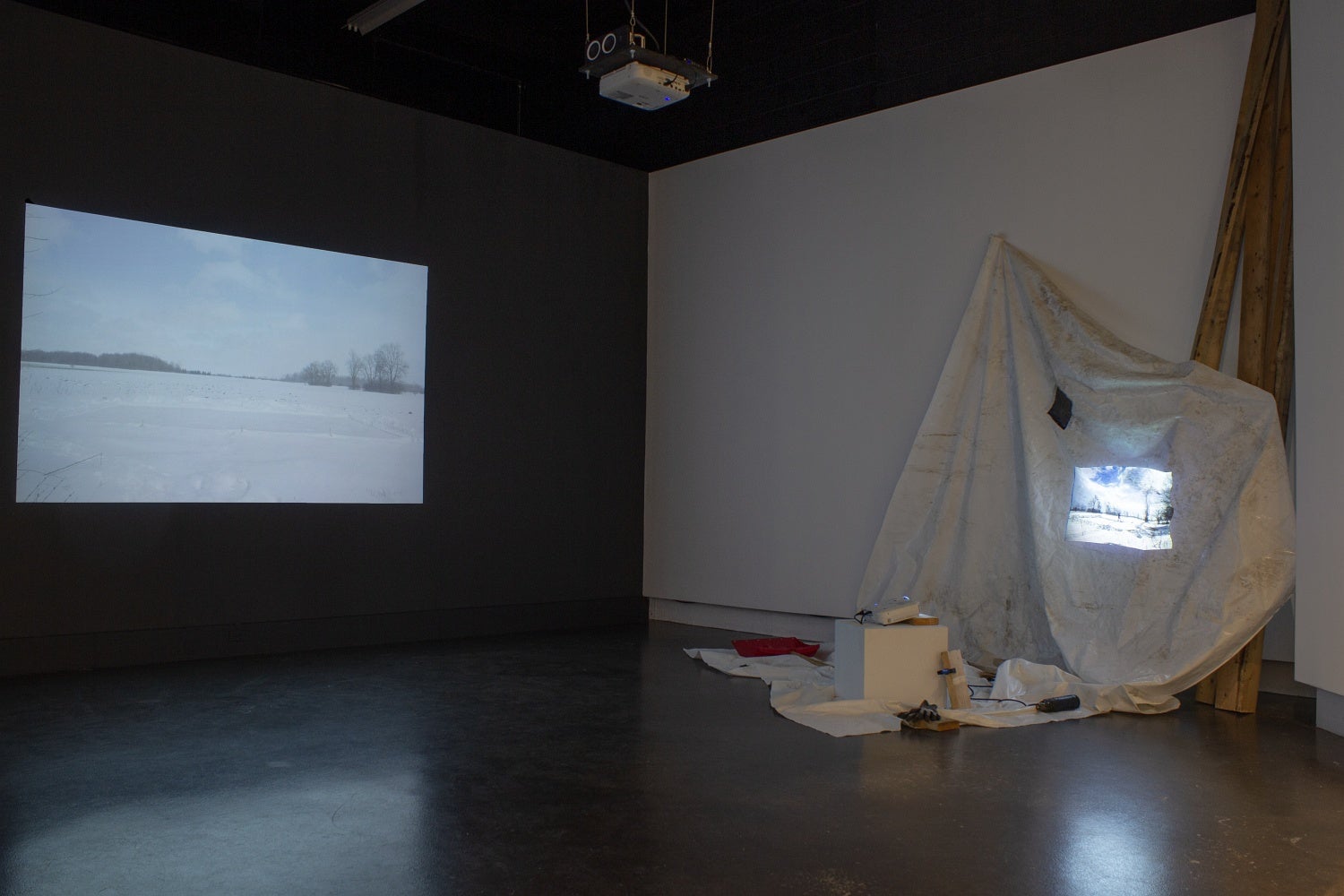 Art gallery with one video projected on a wall and one projected against a crumpled white tarp with various tools and wooden boards piled around the projector