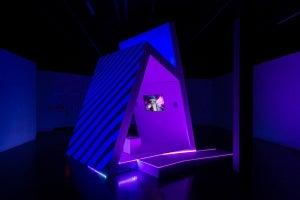 Artwork in gallery.  Triangular structure in room, lighted deep blue and purple