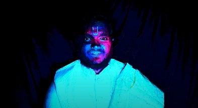 Person wearing a cyan robe is shown from the waist up against a black background.  The face is coloured blue and magenta with a U shaped tilak in the center of the forehead.