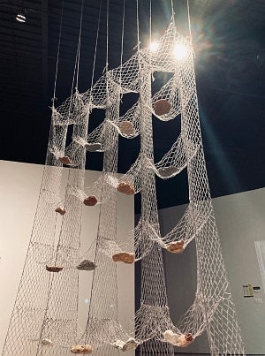 Detail of artwork suspended from a black ceiling. 4-by-4 sections of hammock-like net with each section holding a rock-like object.