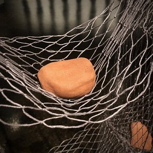 Detail of artwork. Terracotta coloured, rock-like object is suspended in a hammock-like net.
