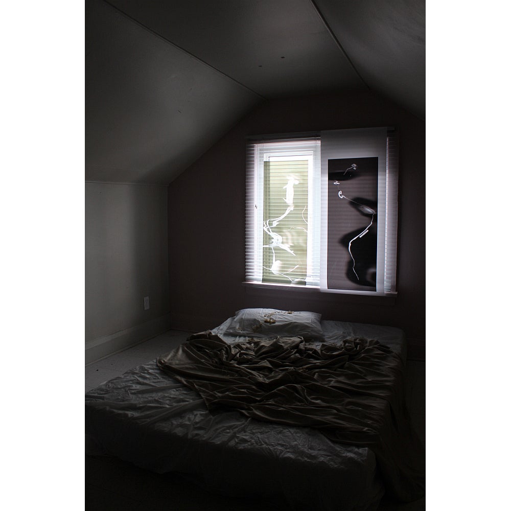Dark room with a mattress and rump led sheets on the floor in front of a window.  Window panes are covered with translucent film