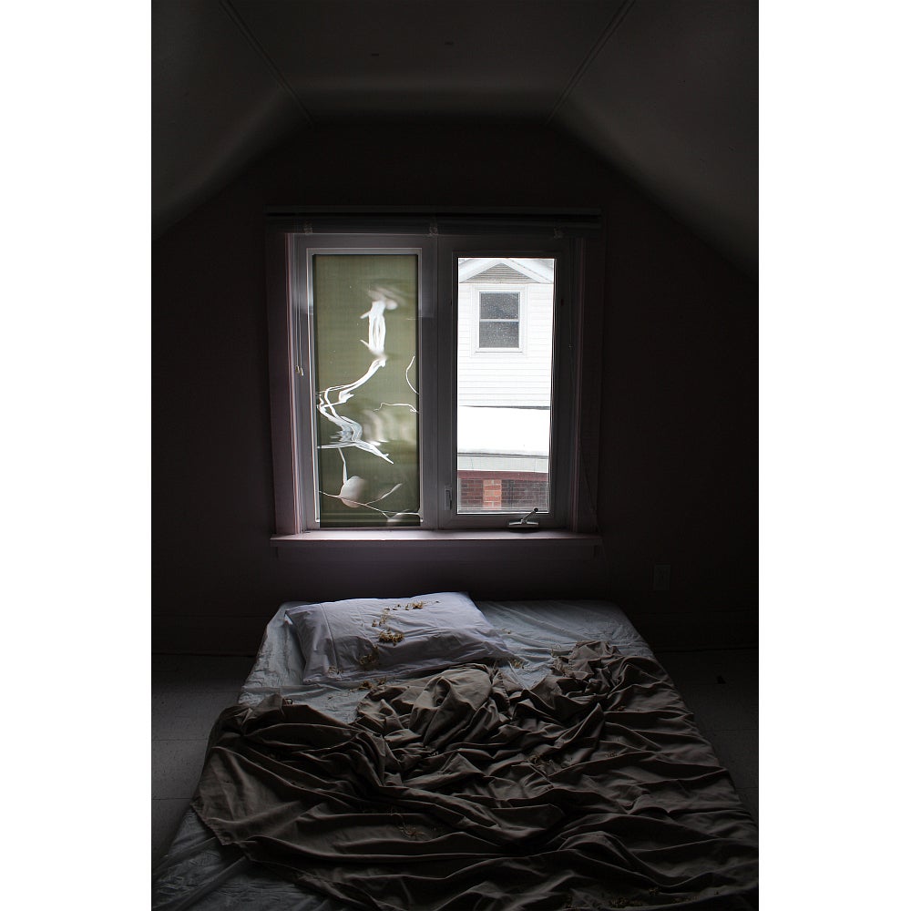 Dark room with a mattress and rumpled sheets on the floor in front of a window.  Window panes are covered with translucent film