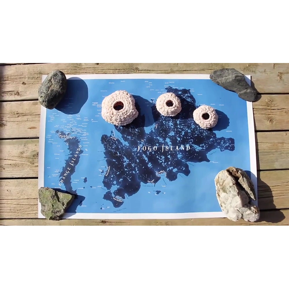 Map of Fogo Island lying flat on wooden deck held down with 4 rocks.  3 small crocheted spheres sit on the map.