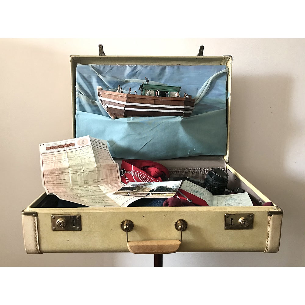 Vintage suitcase opened to reveal document, clothing, camera. Inside top cover contains model of a wooden boat and tiny figures.
