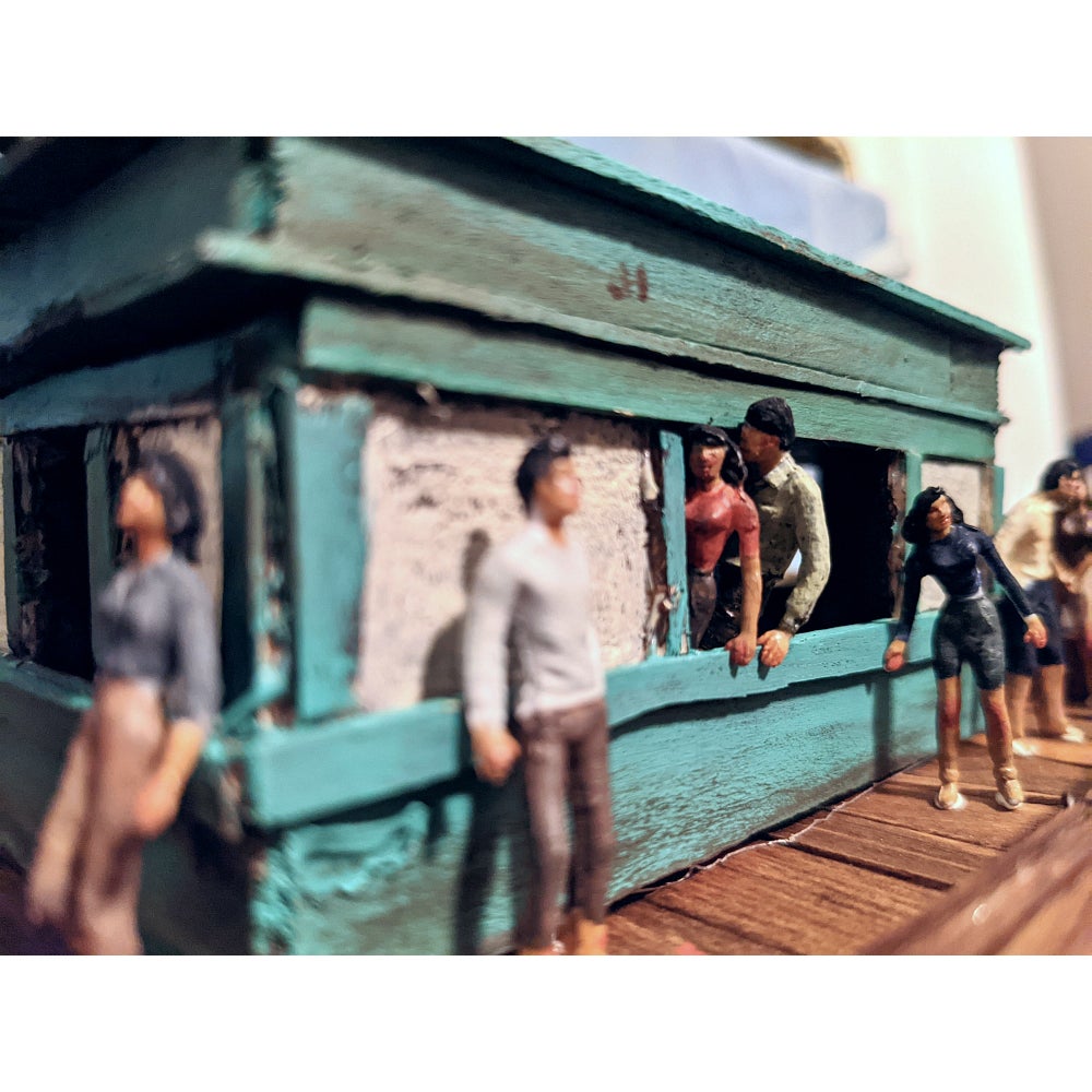 Model of a wooden boat showing deck and teal coloured cabin with tiny figures in various poses.