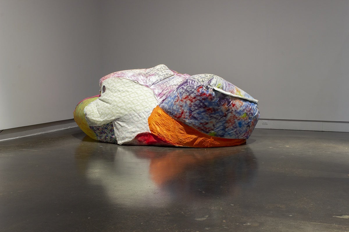 A large, irregularly shaped, inflated sculpture made from various bright fabrics on a gallery floor. 