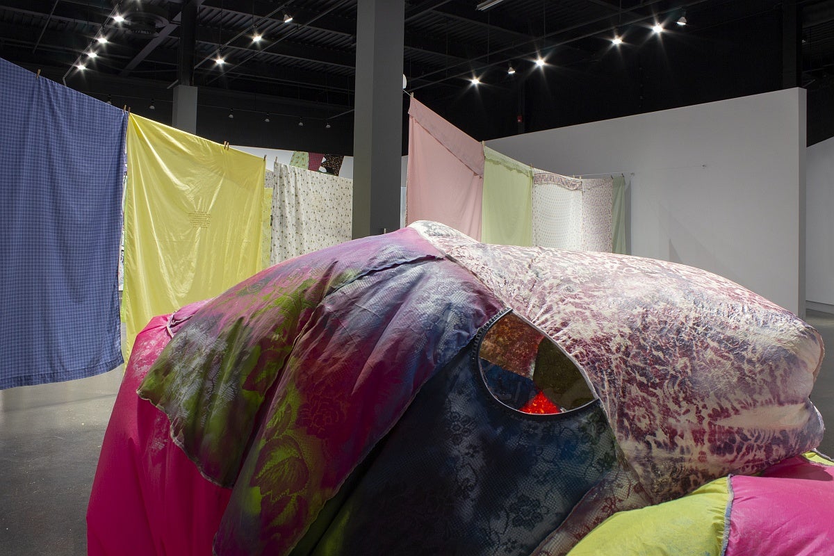 View of an art exhibition with a large, irregularly shaped, inflated sculpture made from various bright fabrics in front of multi-coloured and patterned bedsheets hung on what resemble clotheslines. 