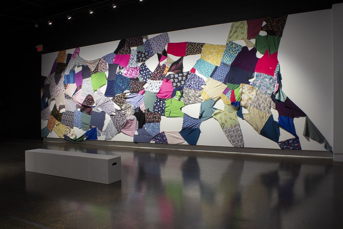An artwork with multi-coloured and patterned caregiver scrubs collaged together in a quilt-like pattern and stretched to cover a wall.
