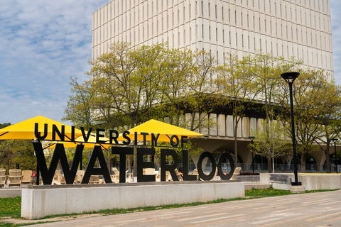 a sign on campus saying University of Waterloo. The main campus library is in the background