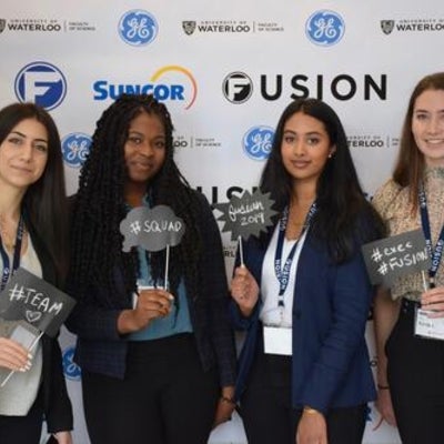 Four females standing in front of Fusion confrence banner