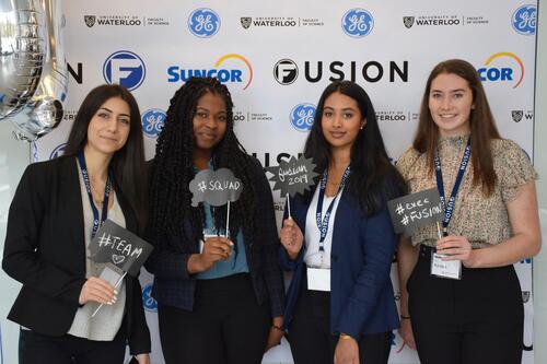 Four females standing in front of Fusion confrence banner