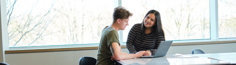 Two students chat in a classroom with a laptop between them