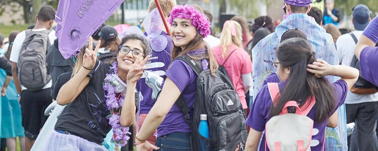 Two students wearing purple and holding purple flag look at the camera