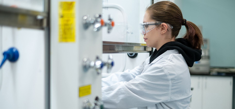 a Medicinal Chemistry student wearing a lab coat and goggles working under a fume hood