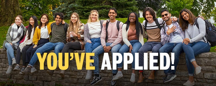 You've applied! Group of students sitting on stone fence smiling at camera.