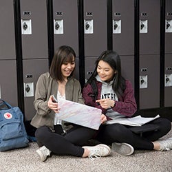 Two students sitting on the floor in front of lockers looking at a laptop together