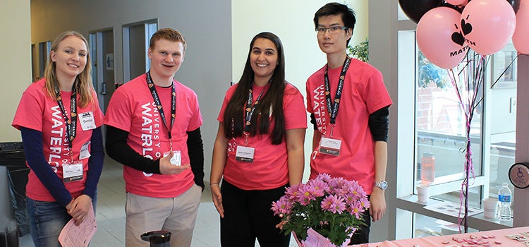 Four Waterloo math students in pink math shirts stand behind a welcome desk