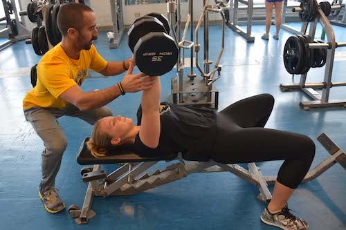 A personal trainer helps a student lift weights
