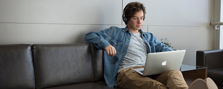 A student sittng on a couch with headphones on and working on a laptop.