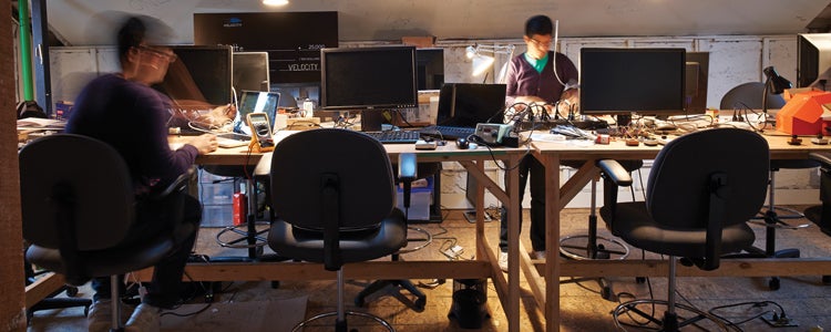 Two students at desk covered in computers and cables in dimly lit room