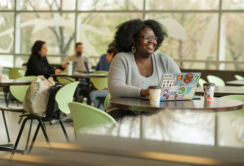Celine Isimbi sits at a table on her laptop in front of a wall of windows