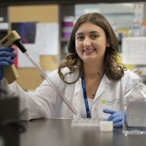 Alessia wears a lab coat and gloves while holding tools in a lab environment