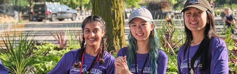 Three students outdoors in nature in purple shirts smiling for the photo.