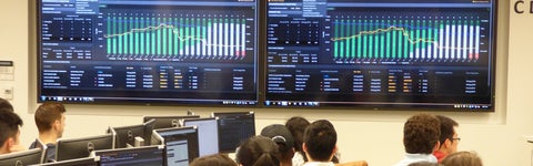 A classroom full of business students looking at two large monitors with statistics and graphs displayed.