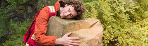 A student in the Earth Sciences degree program hugs a big rock while wearing an orange coat.