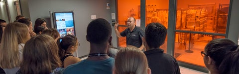 Professor talking to a group of students with a quantum lab in the background