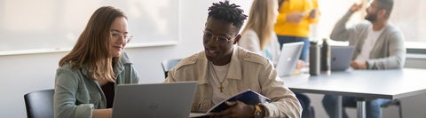 two students sitting in a bright room at a table looking at a laptop