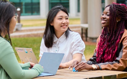 Three students sitting on a table outside on campus with their laptops open