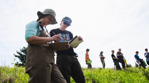 Two students write on clipboards while doing field work