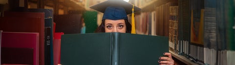 Student wearing a graduation cap, holding a book. Shelves lined with books are on either side of them.
