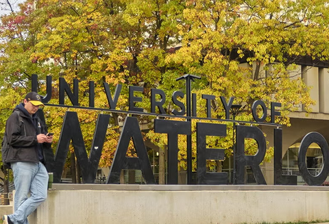 A student on their phone standing in front of the University of Waterloo sign.