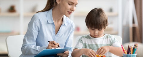 woman holding clipboard and pen looking over at a child doing an activity