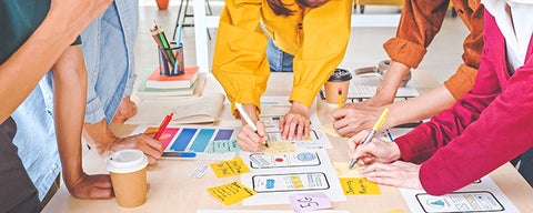 Hands of multiple people working together on sketches and sticky notes spread across a table.