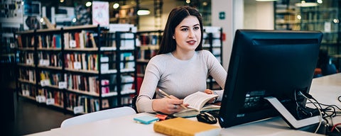 Person sitting at a computer in a library, writing notes whole looking at desktop monitor.