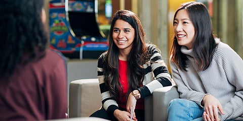 Two students in easy chairs lean forward while talking to another student