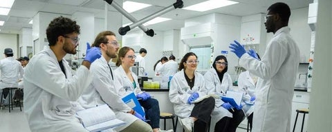Five students in lab coats, gloves, and goggles, sitting infront of an instructor also wearing a lab coat, goggles, and gloves