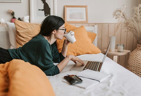 student lying on bed using laptop and drinking from a mug