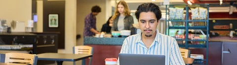 University of Waterloo student looking down at laptop with people buying coffee behind him 