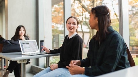 Students chat while seated at tables in front of a window